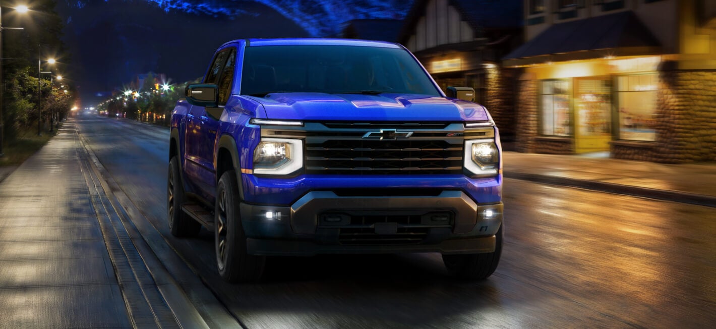 Front exterior view of a blue 2027 Chevy Silverado Z71 driving on a house-lined suburban road,