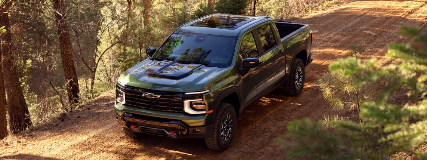 Overhead exterior view of a green 2027 Chevy Silverado Trail Boss driving down a forest dirt road.