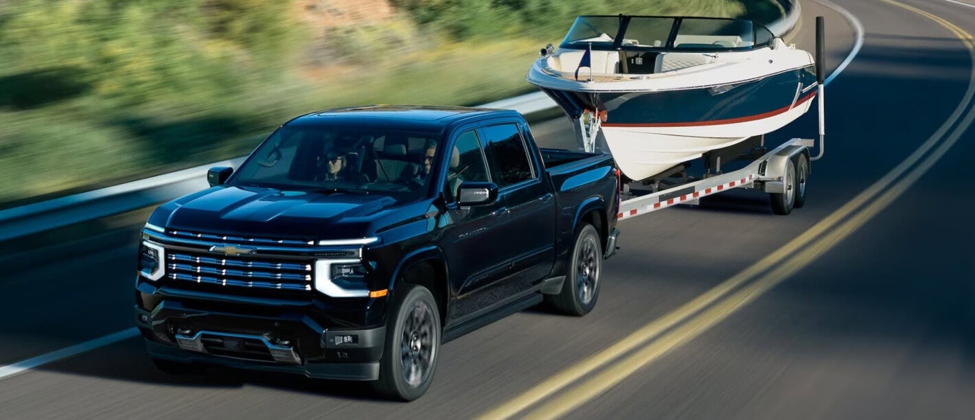 Exterior action shot of a black 2027 Chevy Silverado High Country towing a boat on a highway.