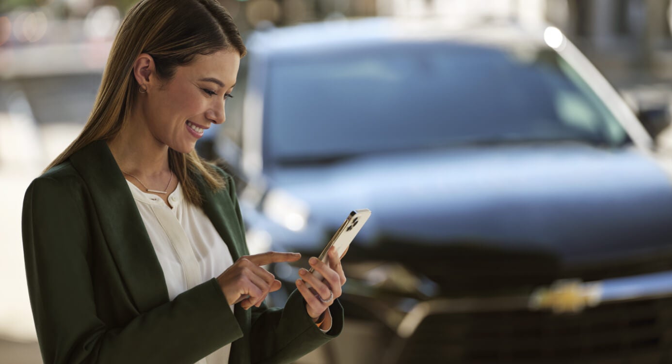 Close-up view of a smiling woman using her smartphone in front of a Chevrolet Blazer on a city street.